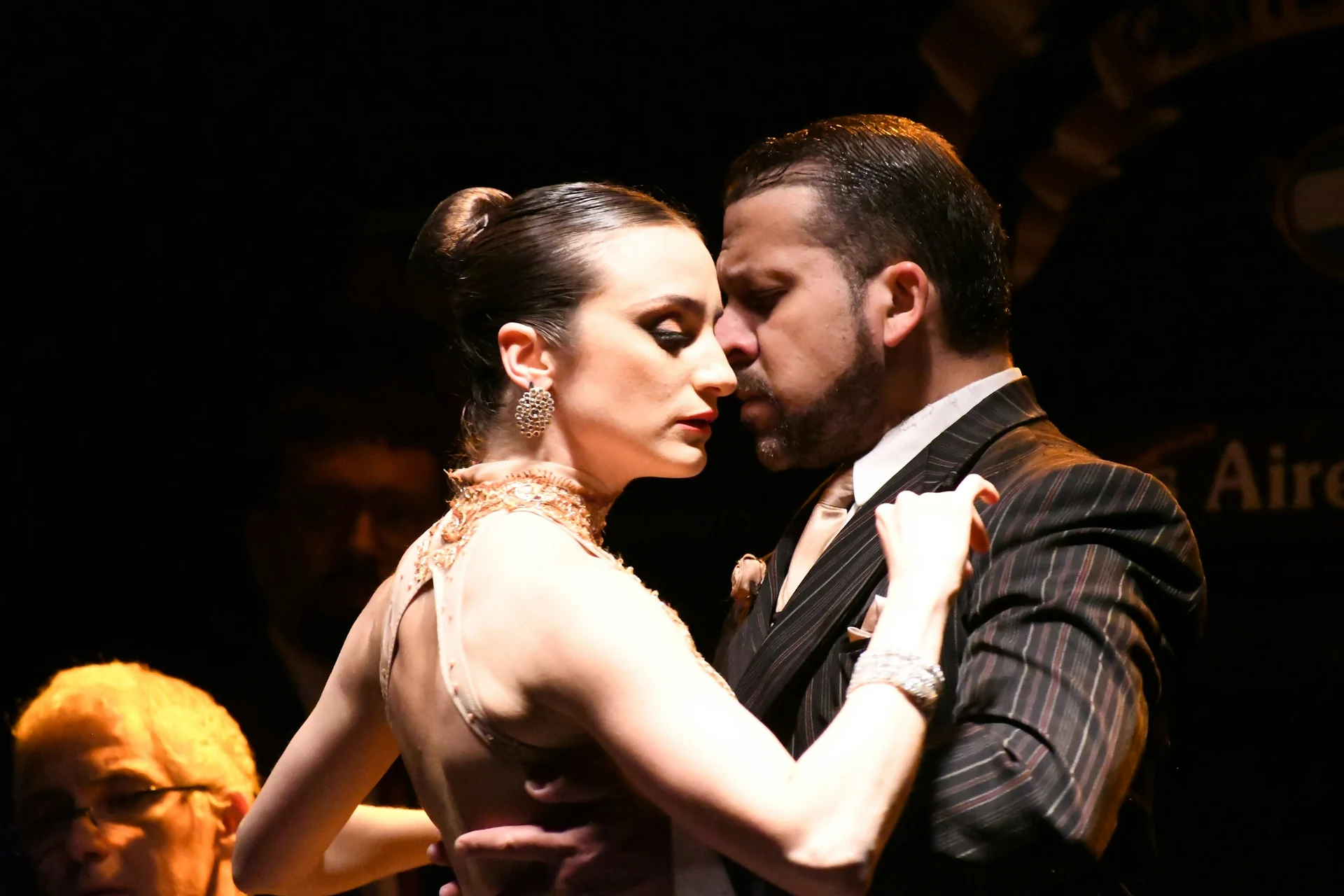 Two tango dancers in formal dress locked in a close embrace on a darkened stage, lit from the side