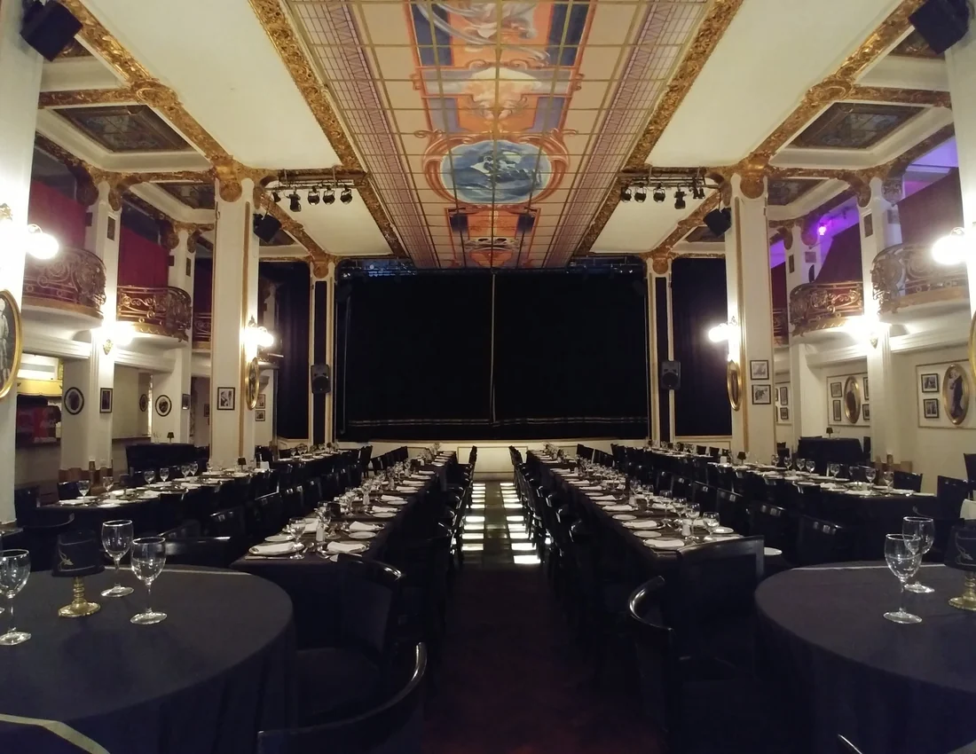 A theatre-restaurant inside the Galeria Guemes set with long dinner tables facing a curtained stage, under a painted glass ceiling